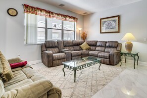 A living room with a sectional sofa, a glass coffee table, a lamp, a clock, and a window with curtains. - Spacious Kissimmee Villa w/ Private Pool & Hot Tub (Kissimmee)