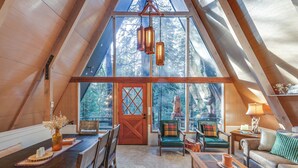 A cozy interior with a wooden ceiling, a dining area with a table and chairs, a sofa, and a large window offering a view of trees. - 3 Mi to Calaveras Big Trees: Cabin w/ Fire Pit! (Arnold)