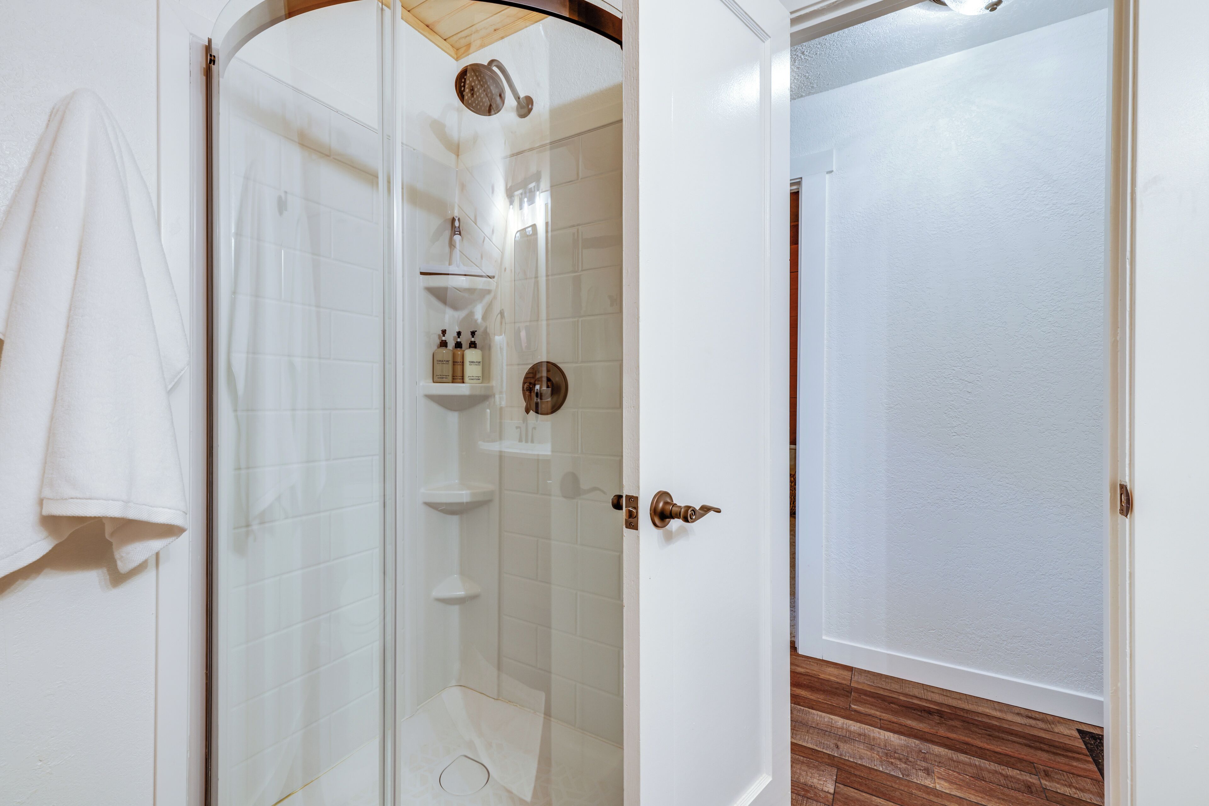 Un baño con ducha envuelta en vidrio, paredes de azulejos blancos, piso de madera y una toalla blanca colgada en la pared.