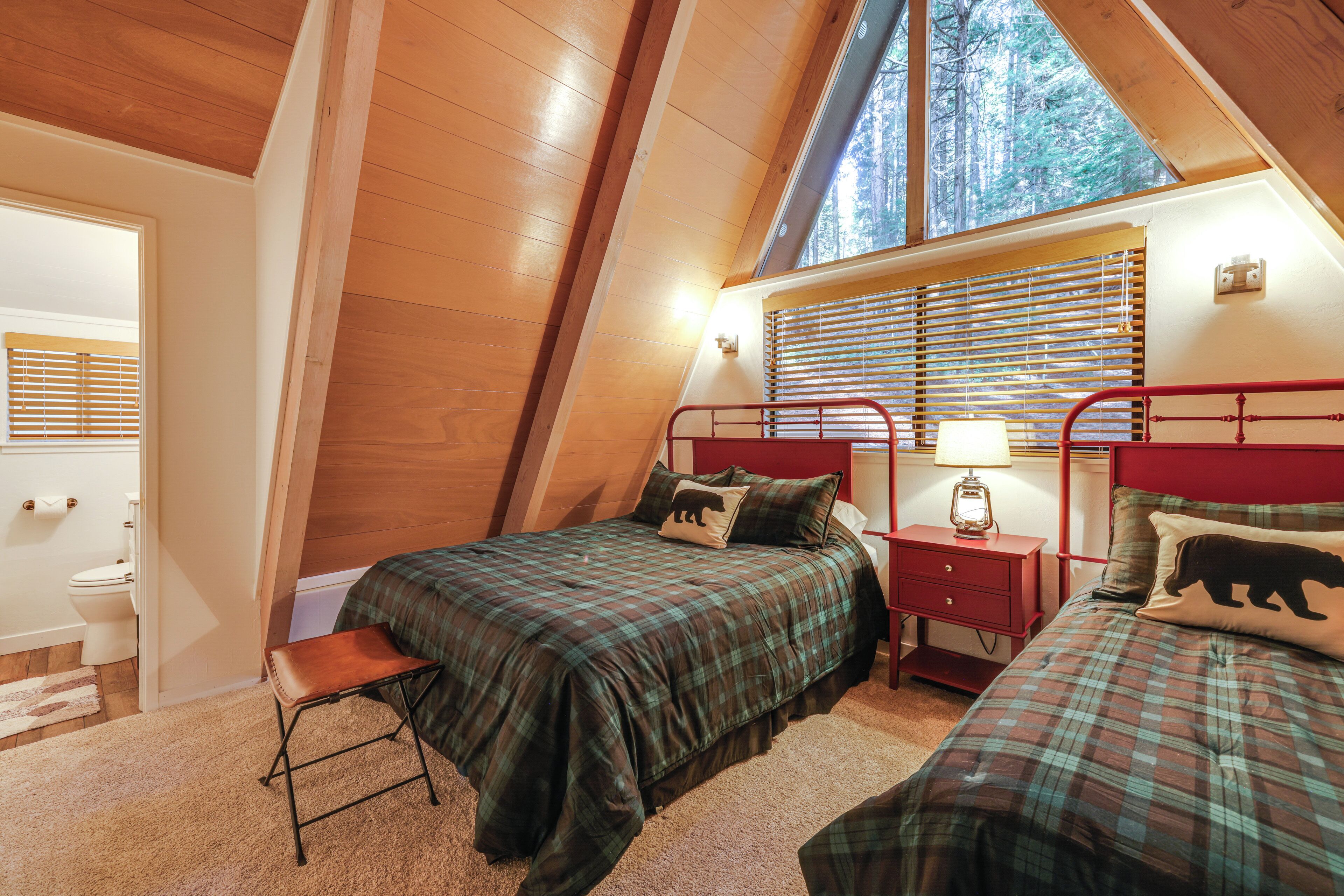Une chambre avec un plafond en pente, un lit recouvert d’une couverture à carreaux, une table de chevet avec une lampe et une fenêtre avec des stores.