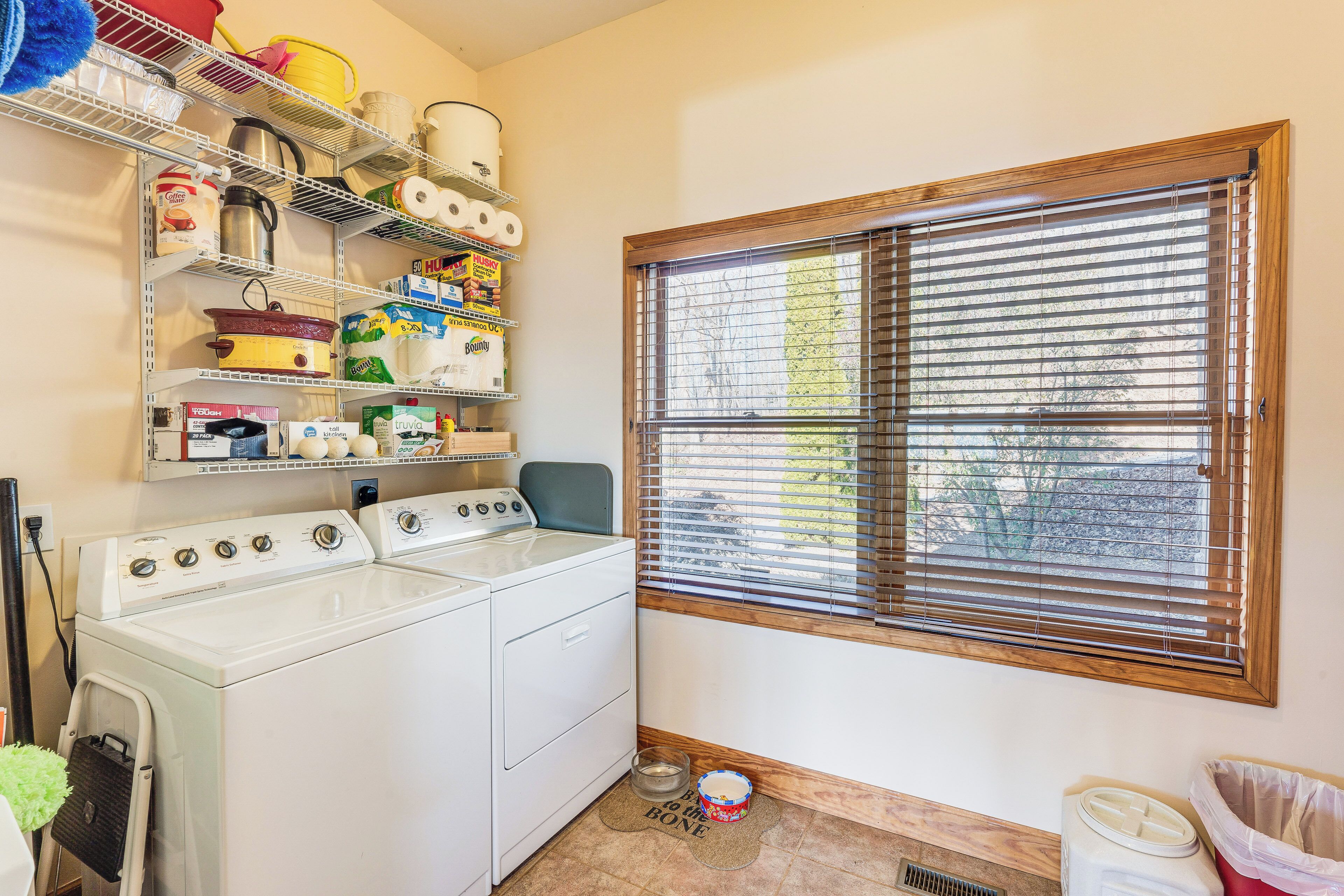 A laundry room with a washer and dryer, shelves with various household items, a window with blinds, and a small rug on the floor.