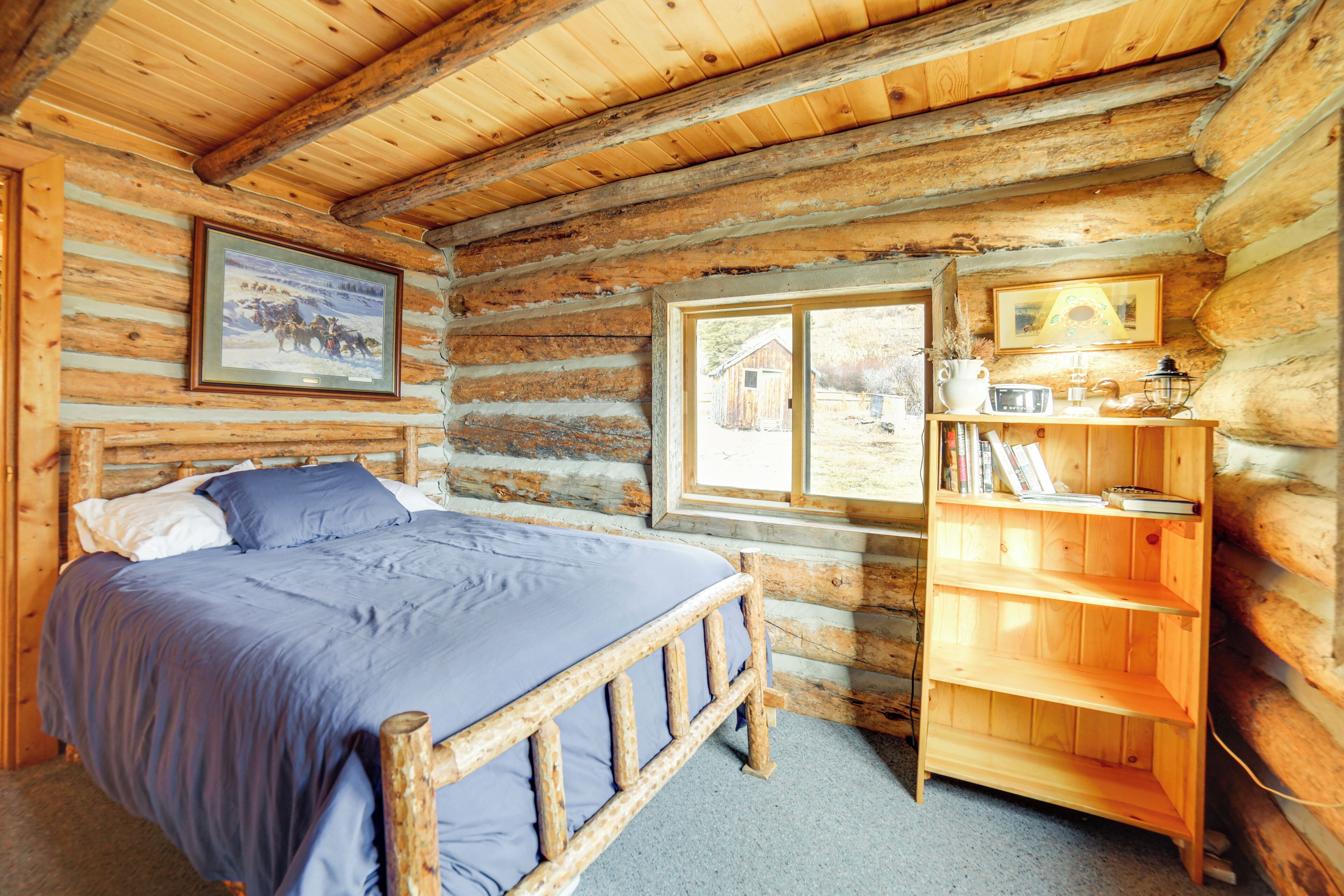 A log cabin bedroom with a bed, a wooden bookshelf, and a window with a view of the outside.