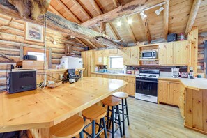 Interior rústico de cabana, paredes de madeira, uma grande mesa de jantar de madeira com banquetas altas, cozinha com fogão, micro-ondas e armários.