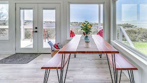 Un coin repas avec une table et des bancs en bois, un vase rempli de fleurs et une vue sur l’extérieur grâce à de grandes fenêtres.