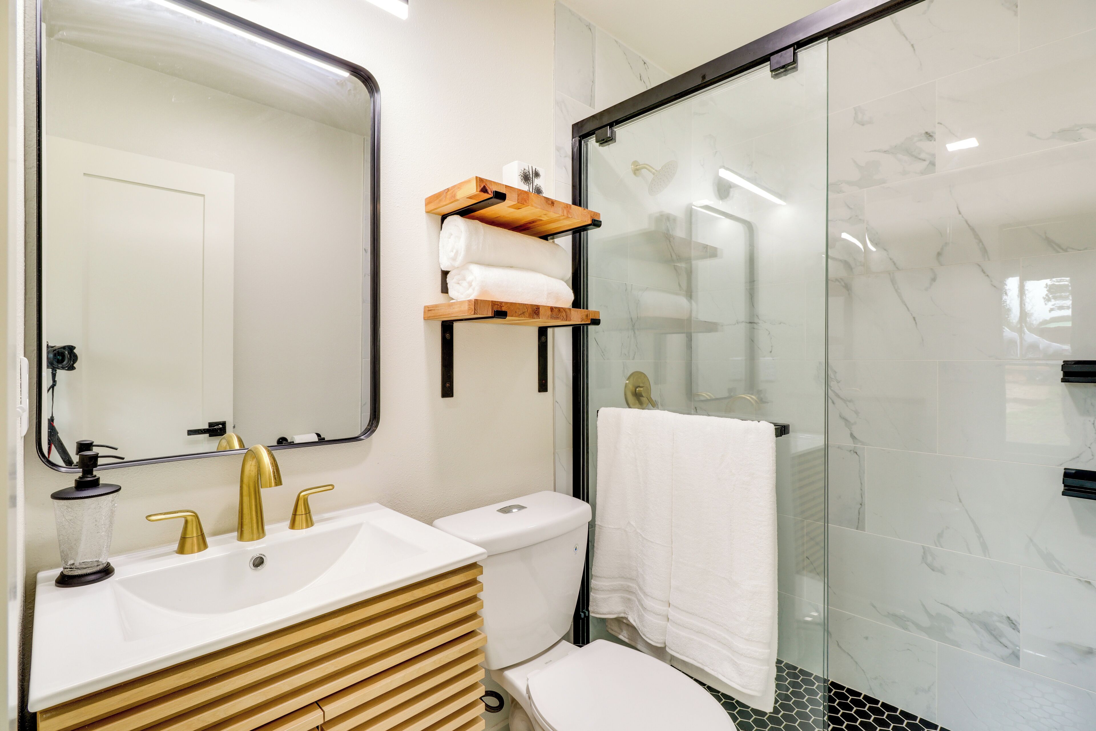 A modern bathroom with a white sink, gold faucets, a toilet, and a glass shower enclosure.