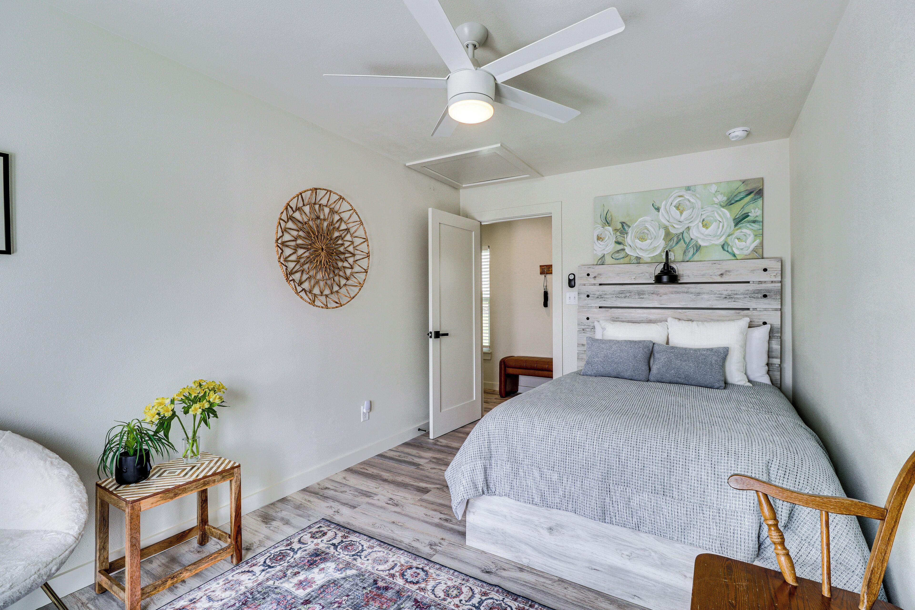 Une chambre à coucher avec un lit, une table de chevet en bois ornée de fleurs, un ventilateur de plafond et un tableau accroché au mur.