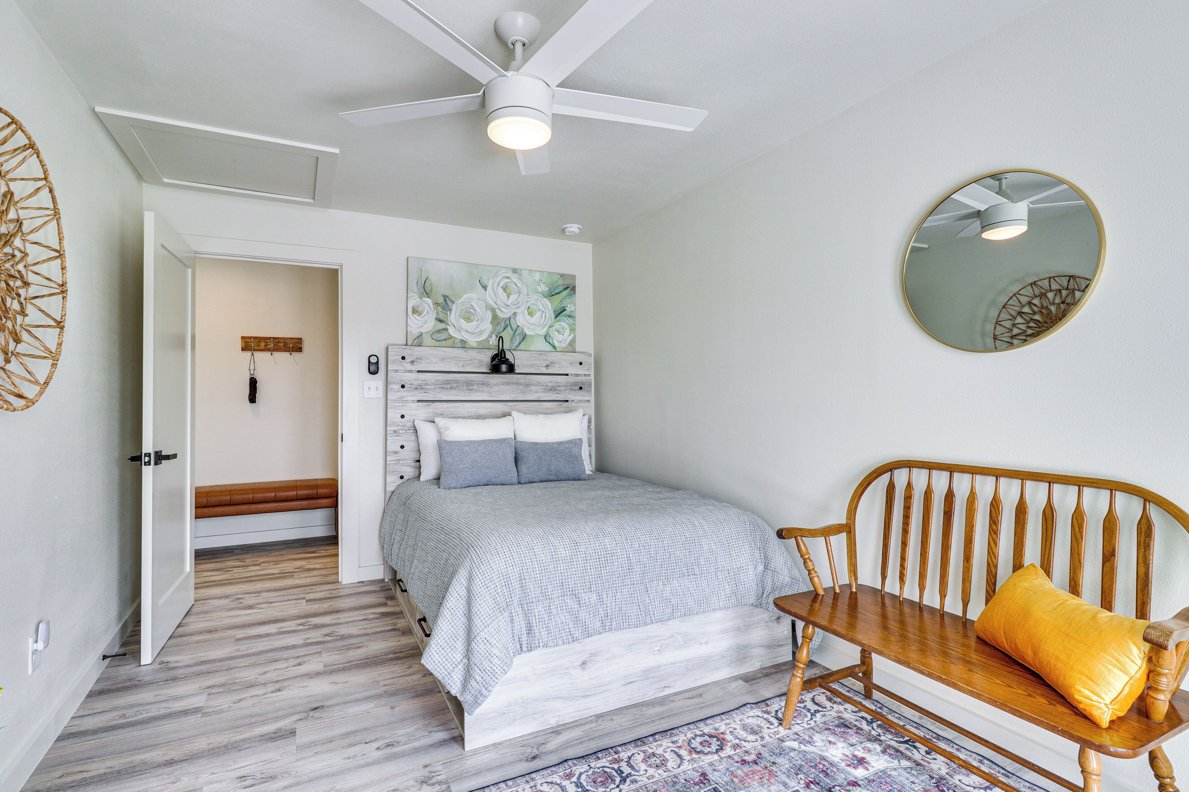 A bedroom with a bed, a wooden bench, a ceiling fan, a mirror, and a door leading to another room.