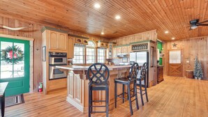 A wooden cabin interior with a kitchen area featuring a central island with bar stools, wooden cabinets, and a refrigerator. - Countryside Cabin Rental ~ 10 Mi to Wadesboro! (Wadesboro)