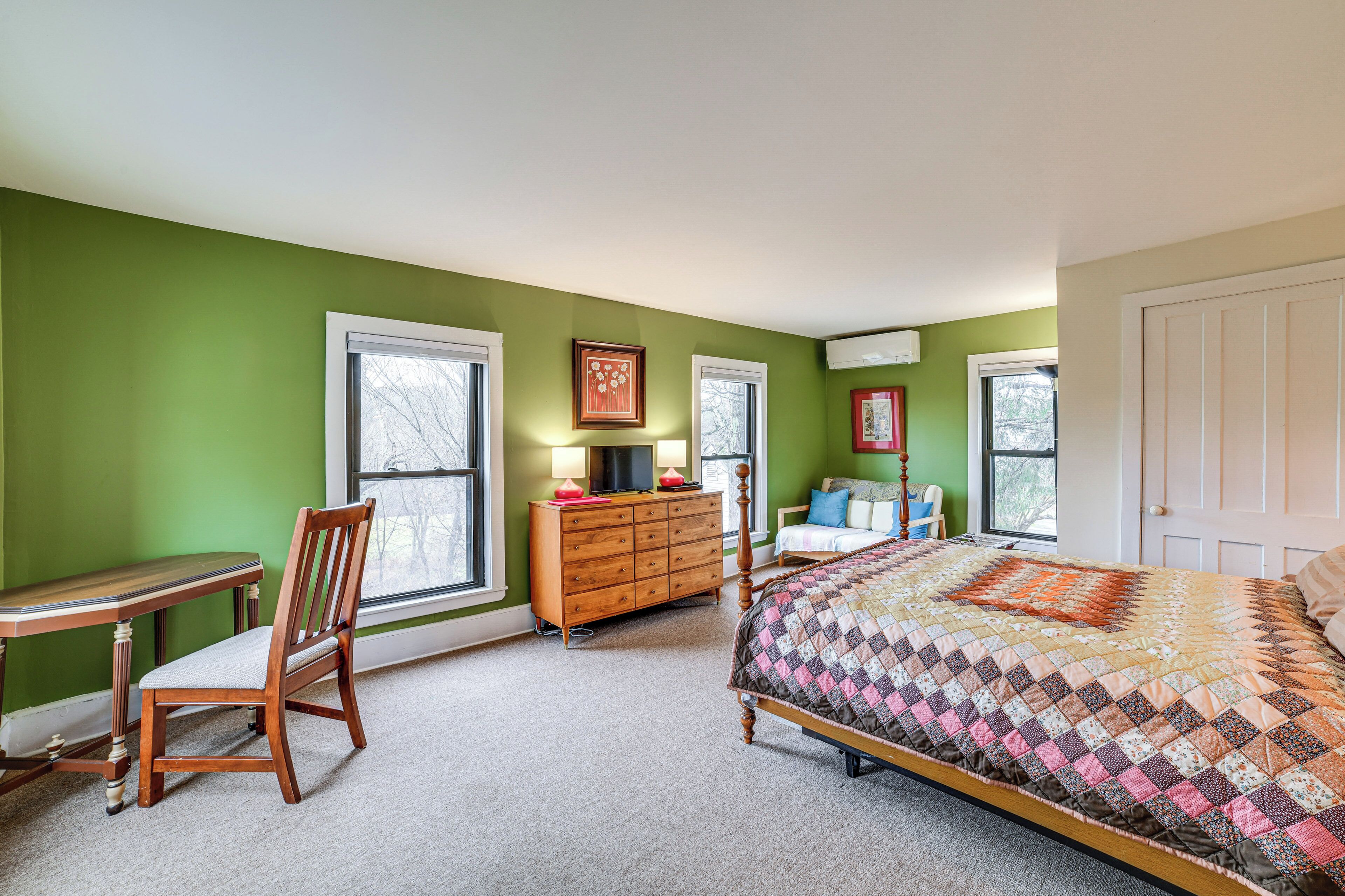 Une chambre avec des murs verts, une commode en bois, un lit recouvert d’une courtepointe à motifs et une chaise.