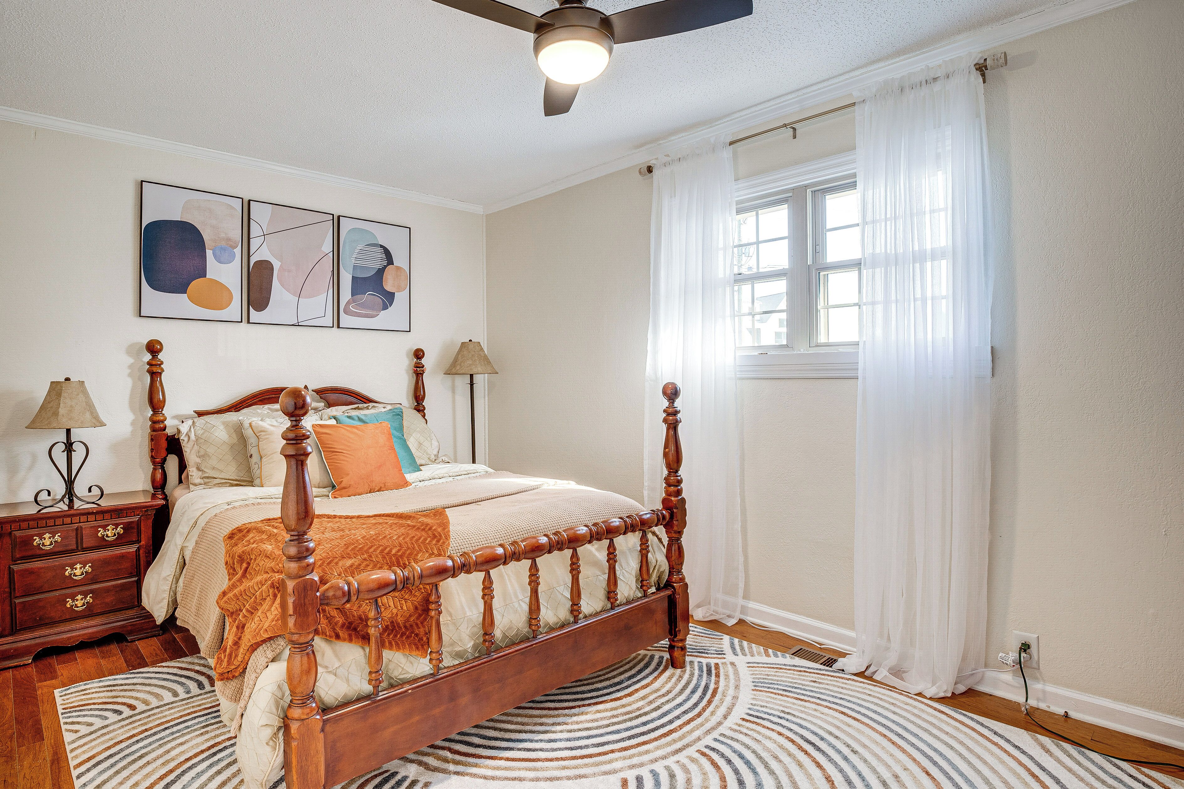 A bedroom with a wooden four-poster bed, a round rug, a ceiling fan, and a window with sheer curtains.