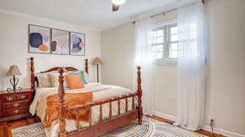 A bedroom with a wooden four-poster bed, a round rug, a ceiling fan, and a window with sheer curtains.