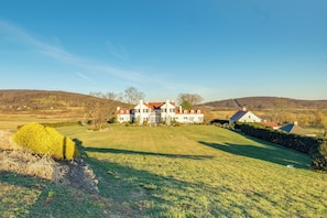 Une grande maison blanche, surmontée d’un toit rouge, entourée d’une pelouse bien entretenue et avec, en arrière-plan, un paysage vallonné.