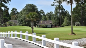 A wooden bridge with white railings leading to a golf course with a clubhouse in the background. - 2804 2 Bedroom Villa by RedAwning (Calabash)