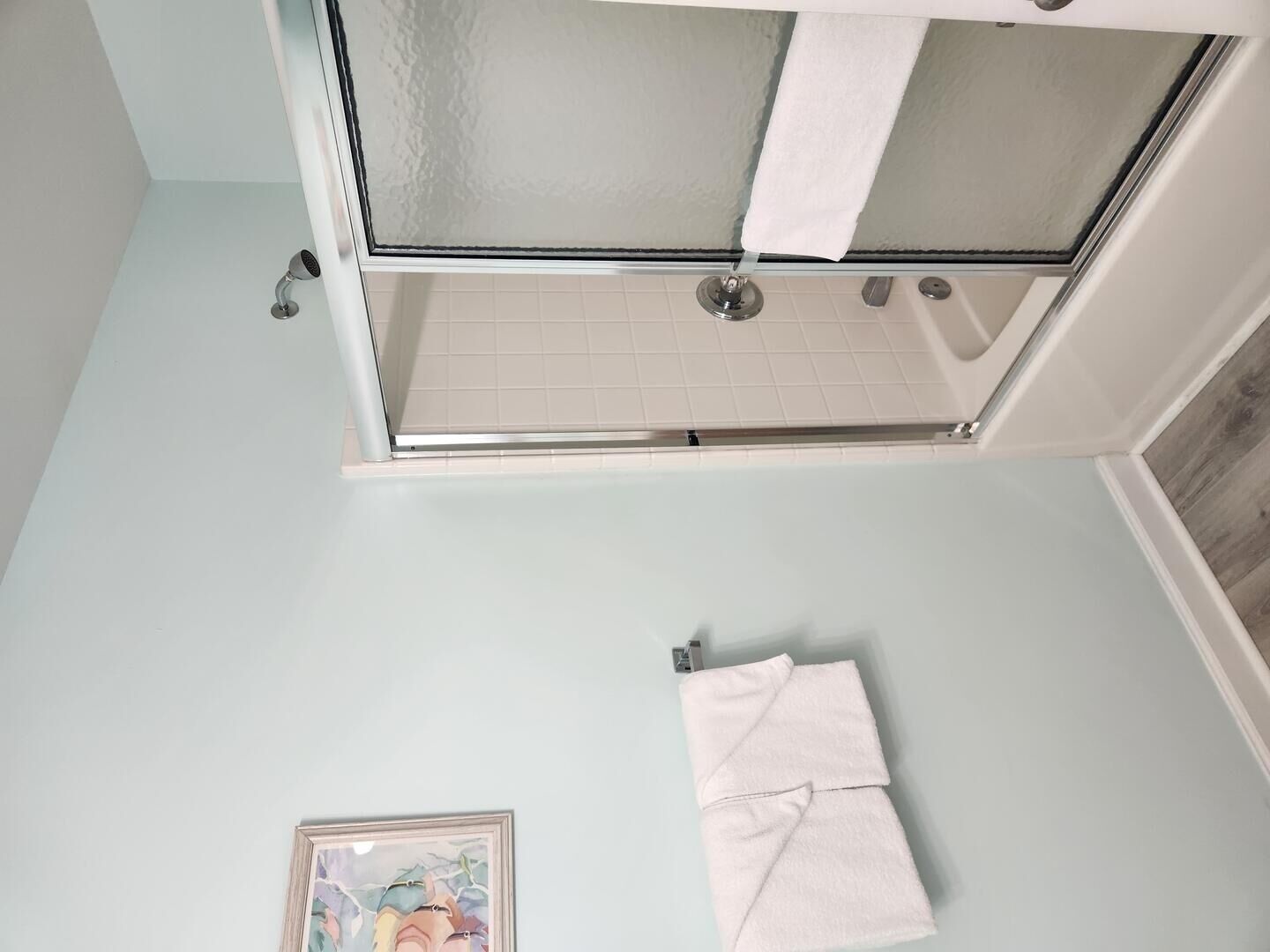 A bathroom with a glass shower enclosure, a white towel hanging on the wall, and a framed picture.