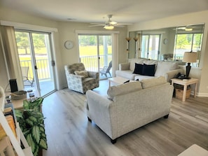 A spacious living room with a sofa, armchair, and a small table. There is a ceiling fan, a large window, and a view of a balcony.