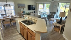 A modern kitchen with a central island, a dining area with wooden chairs, a living room with a sofa and a TV, and a balcony with outdoor furniture.