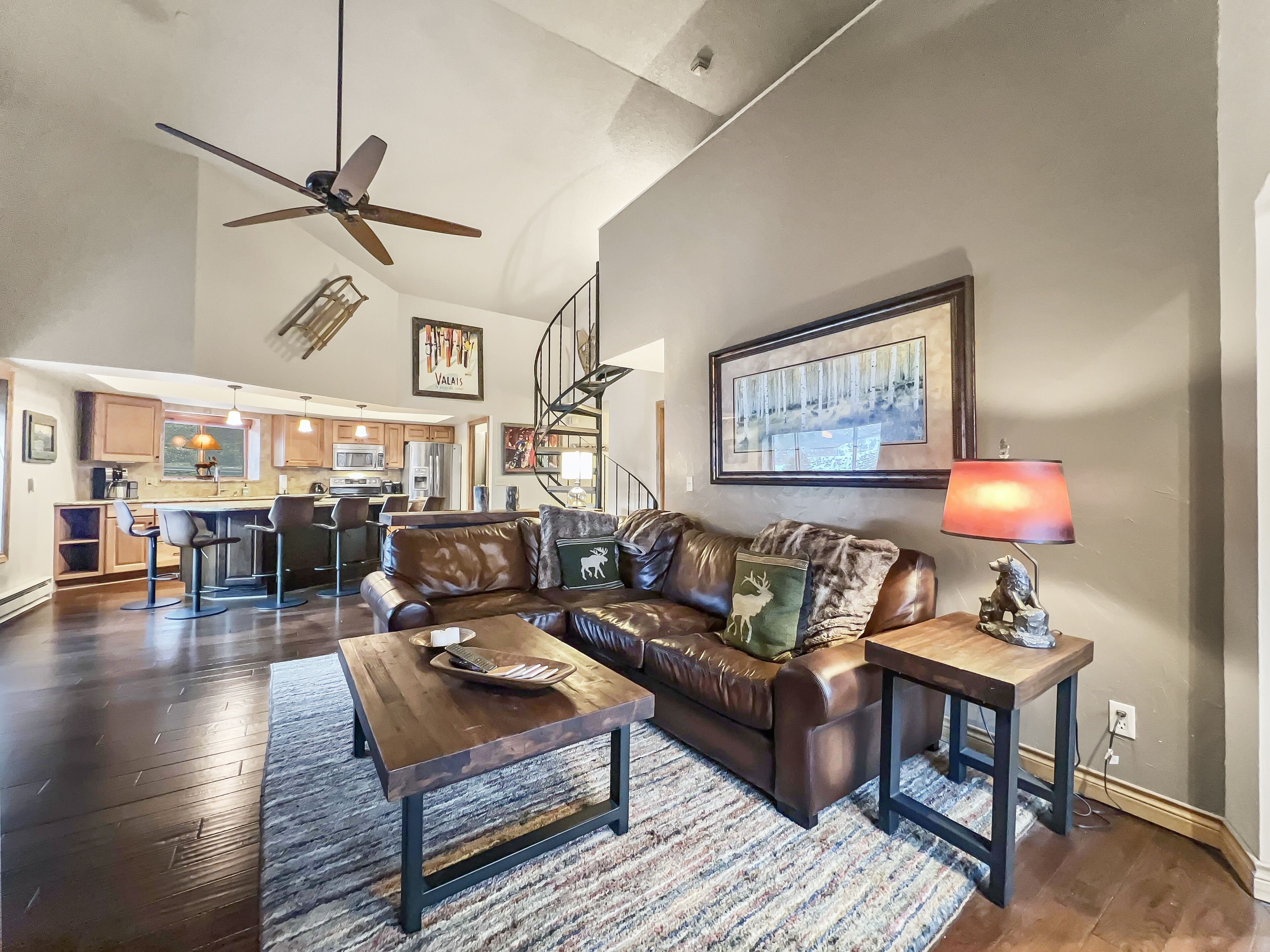 A spacious living room with a leather sofa, a coffee table, and a dining area with a bar. A staircase leads to an upper level, and a ceiling fan is visible.