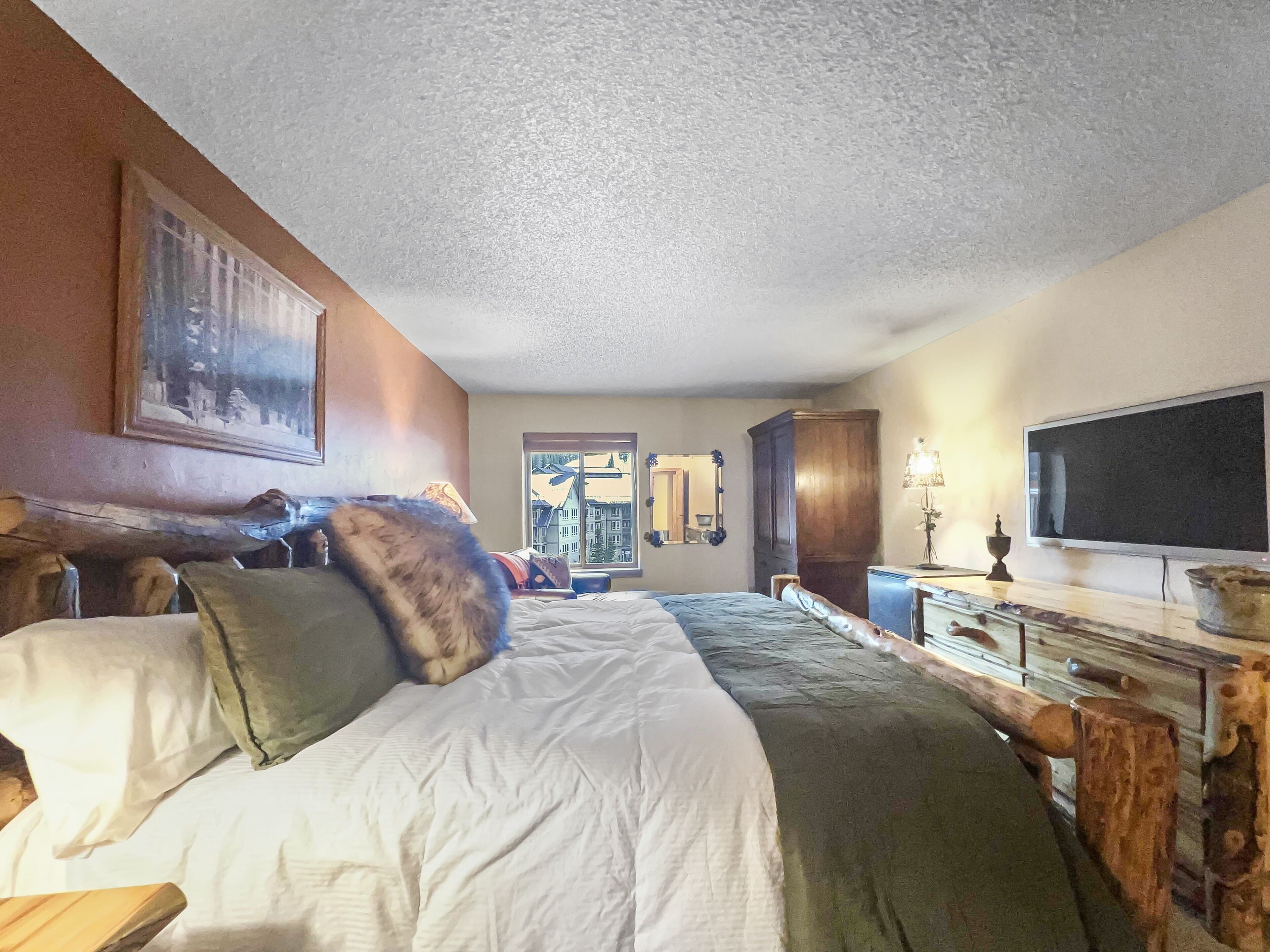 A hotel room with a large bed, a wooden dresser, a television, and a window with a view of buildings.