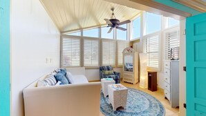 A room with a white sofa, a patterned armchair, a small table, and a blue patterned rug. There is a ceiling fan, a window with blinds, and a door with a turquoise frame. - Augusta Antics 530 ~Golf Cart Included~Pet Friendly~Sandestin Resort (Miramar Beach)