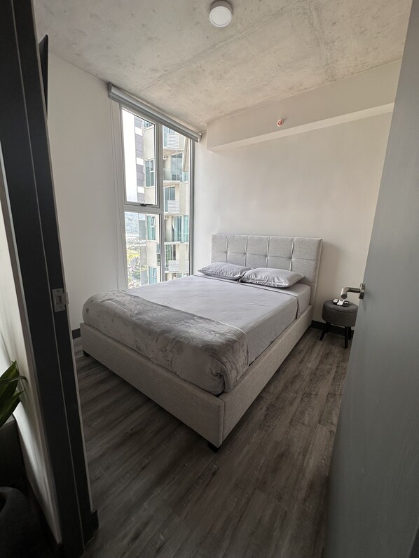 Un dormitorio moderno con una cama grande, una ventana y una mesita de noche pequeña.