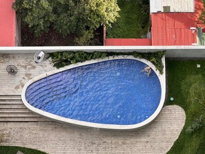 Una piscina con cubierta de madera, rodeada de vegetación y un edificio rojo al fondo.