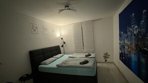 Un dormitorio con una cama, un ventilador de techo, una mesita de noche, una silla, una ventana con persianas y una pintura de paisaje urbano en la pared.