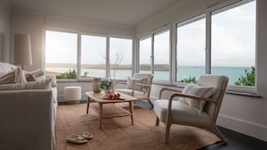 A spacious living room with a large window offering a view of the sea. There is a sofa, a chair, and a coffee table with a plant and fruit on it. - Tregirls - Polzeath -  ocean view, cliff top property sleeping 10 (Polzeath)