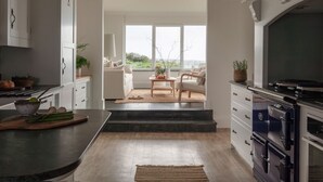 A modern kitchen with a large island, built-in oven, and a view to a living area with a sofa and table. - Tregirls - Polzeath -  ocean view, cliff top property sleeping 10 (Polzeath)