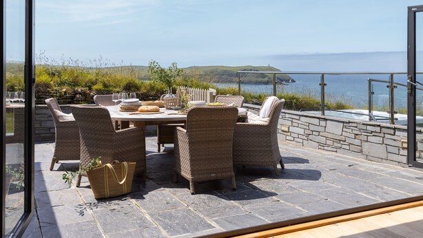 An outdoor dining area with wicker chairs and a table set for four, overlooking a body of water and hills. - Tregirls - Polzeath -  ocean view, cliff top property sleeping 10 (Polzeath)