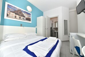 Een net opgemaakt bed met witte lakens en een blauw accent, een klein bureau met stoel, een badkamer met wastafel en spiegel, en een ingelijst schilderij aan de muur.