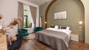 A modern bedroom with a large bed, a green chair, a wooden nightstand, and a window with curtains. - Haller Boutique (Targu Mures)