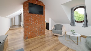 A modern loft with a brick fireplace, a television mounted on the wall, a cozy seating area with a chair and a small table, and a large window with curtains. - Haller Boutique (Targu Mures)