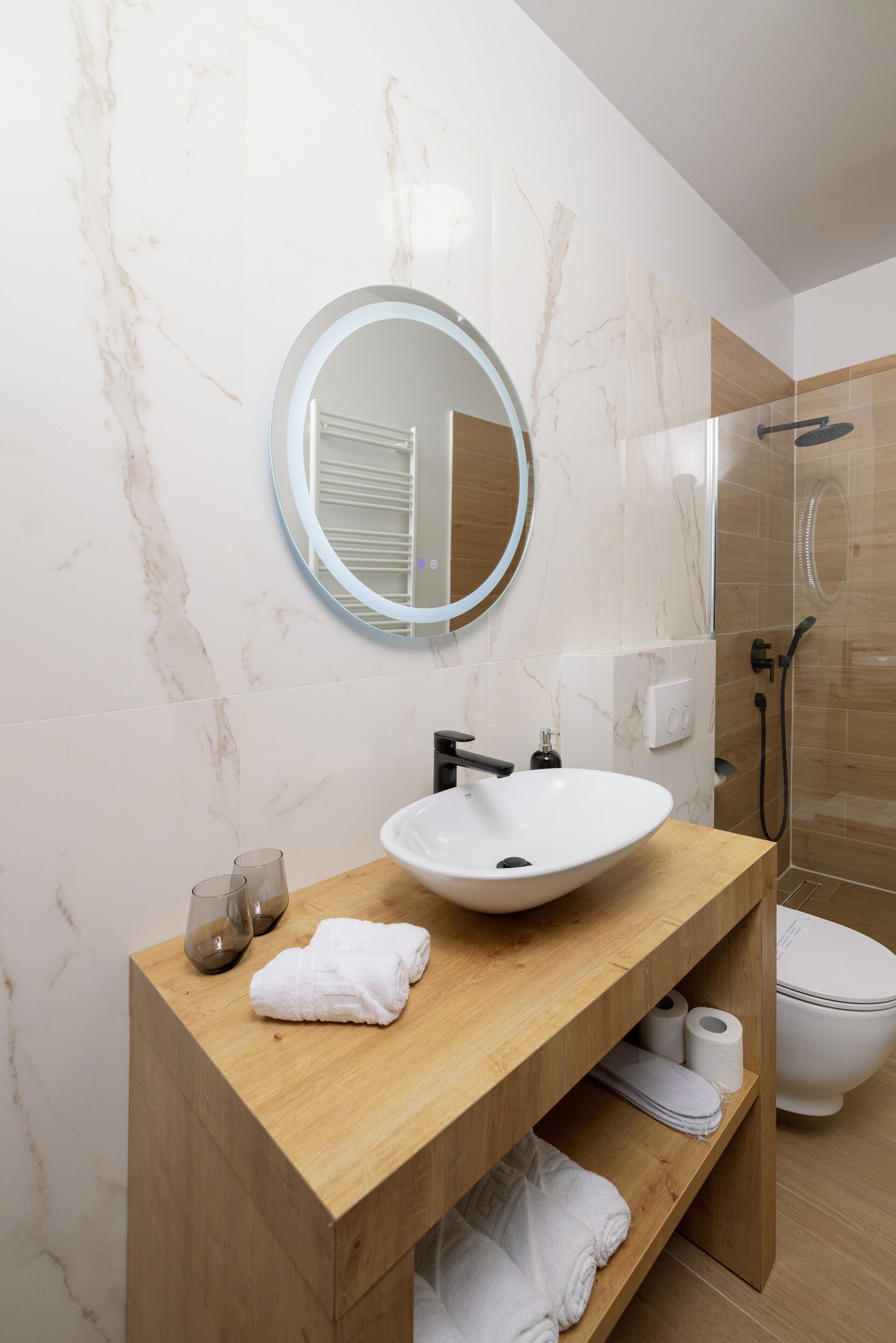 Um banheiro moderno com parede de mármore, espelho redondo, toucador de madeira, pia branca e chuveiro com divisória de vidro.