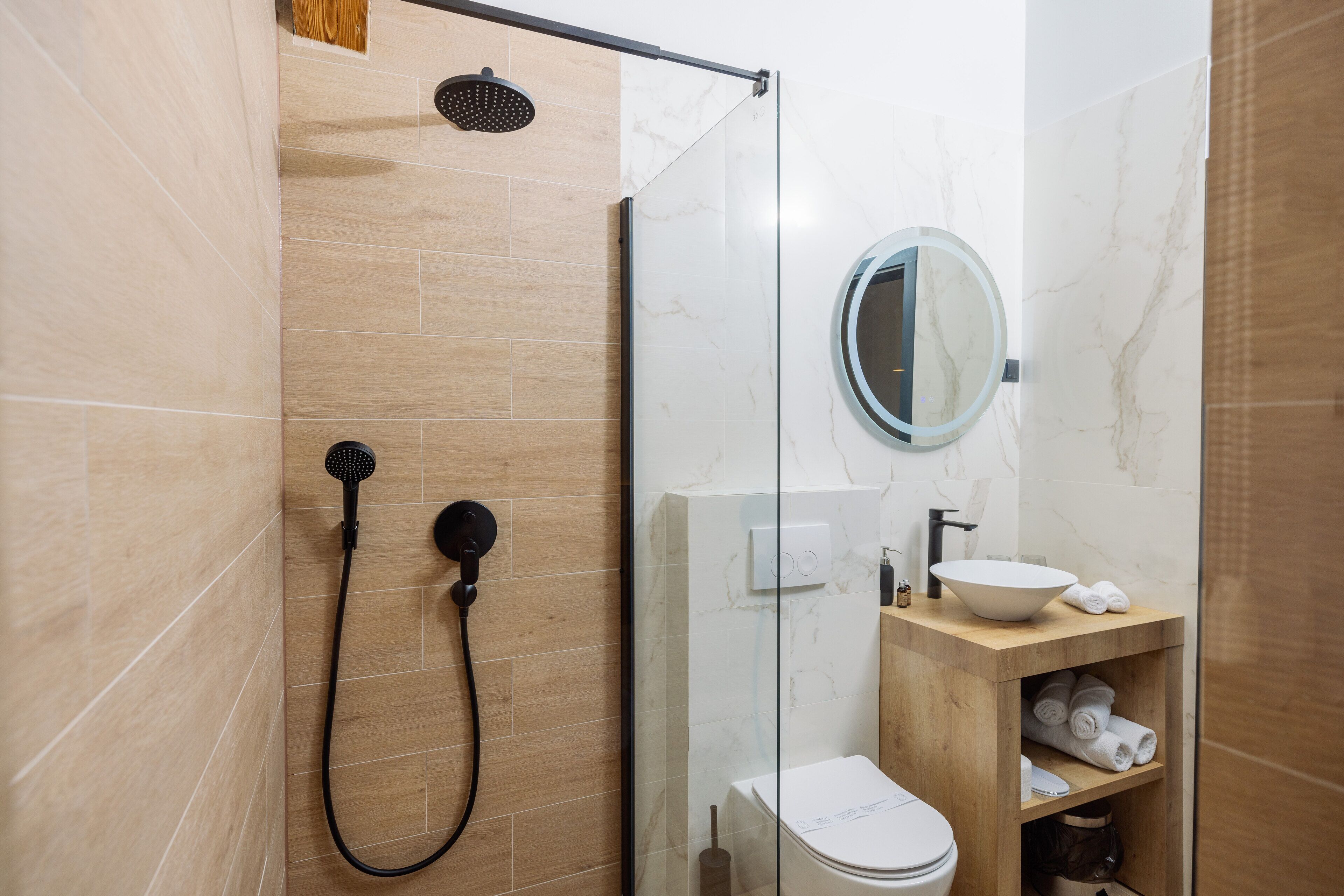 A modern bathroom with a glass shower enclosure, a toilet, a sink with a mirror, and a wooden vanity with towels.