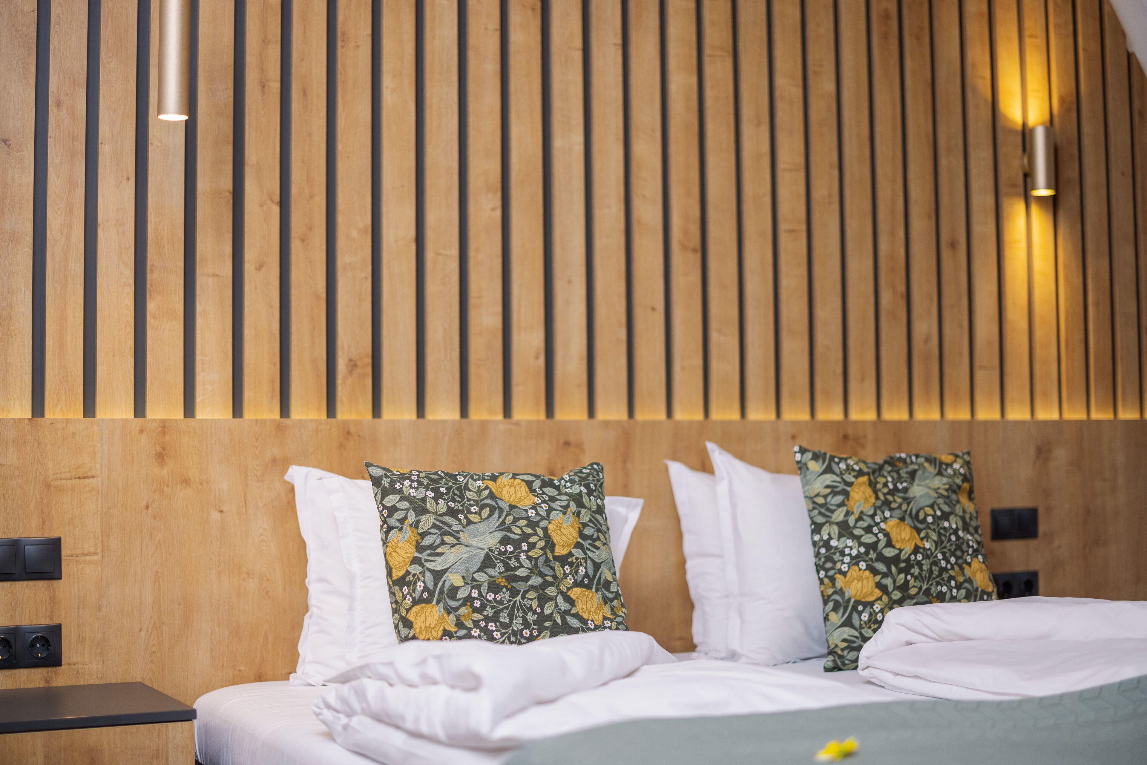 A hotel room with a wooden slat wall, a bed with white bedding, and two decorative pillows with floral patterns.