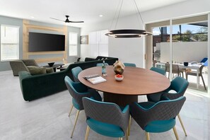 A modern living room with a round wooden table, teal chairs, a green sofa, and a flat-screen TV mounted on the wall.