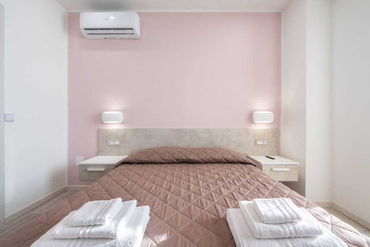 Un dormitorio moderno con pared rosa, cabecero color beige, colcha acolchada, dos mesitas de noche con lámparas y aire acondicionado instalado en el techo.