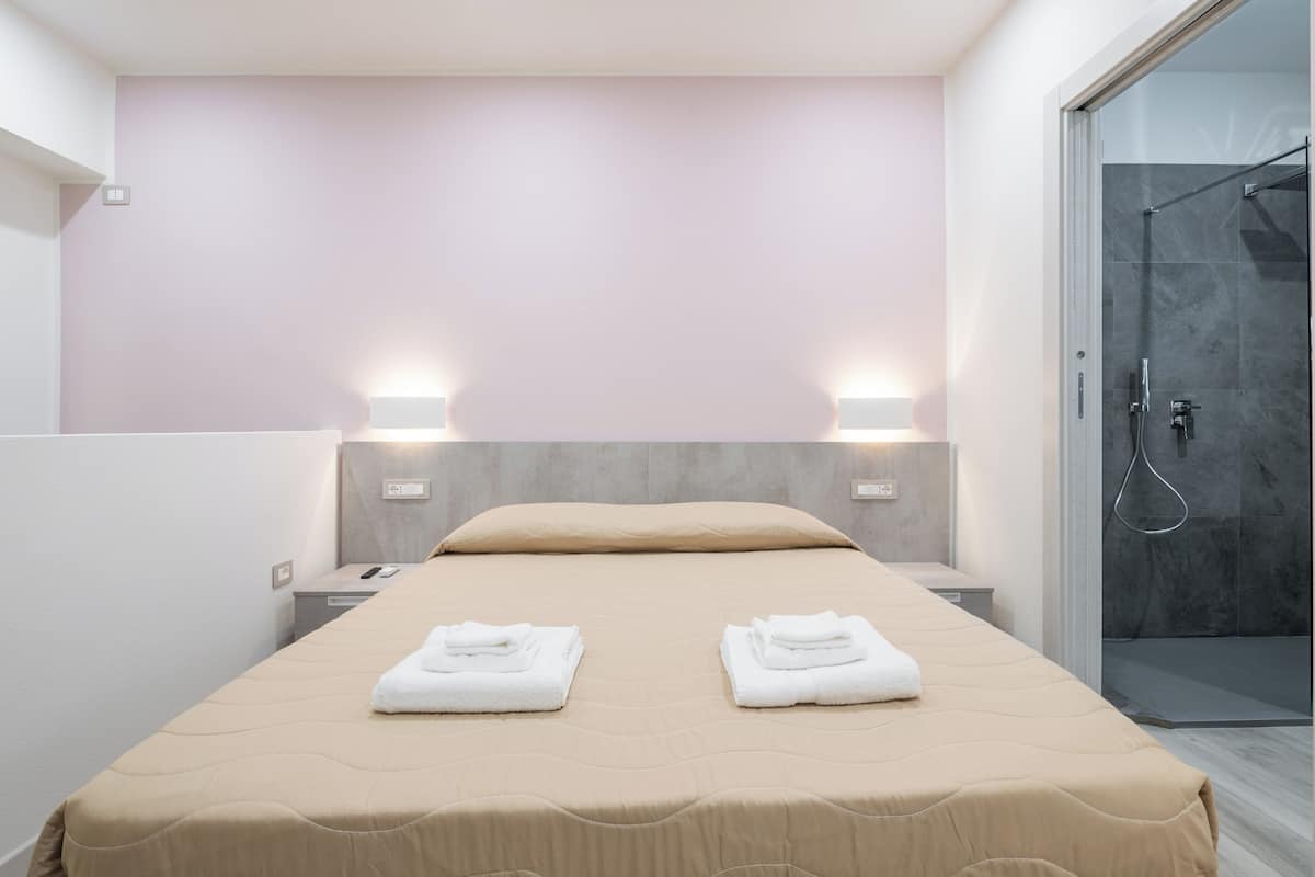 Una habitación de hotel moderna con una cama, dos mesitas de noche, una lámpara de noche, cabina de ducha de vidrio y luces empotradas en la pared.