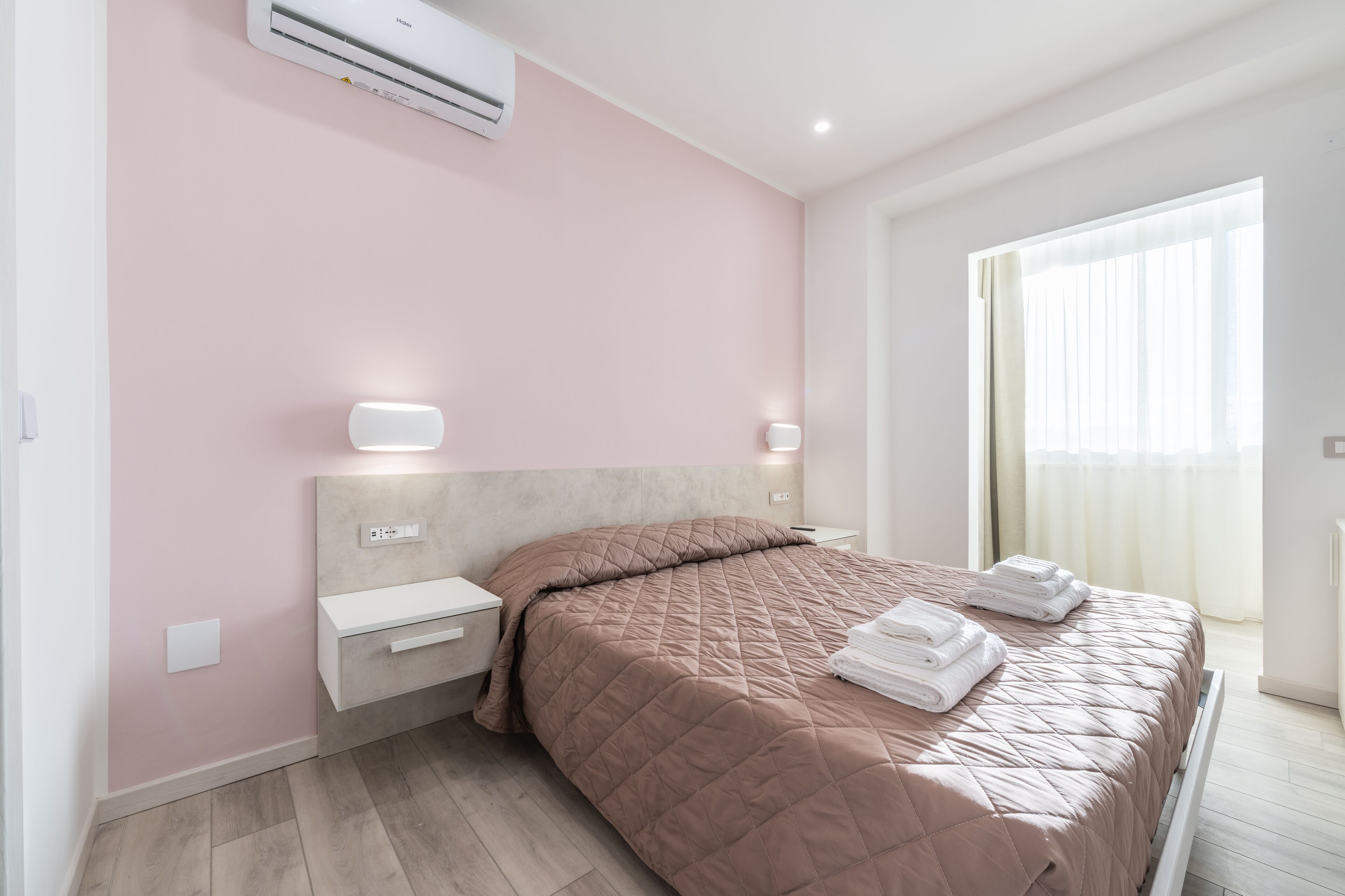 A modern bedroom with a pink wall, a bed with a quilted headboard, bedside tables with lamps, and a large window with curtains.