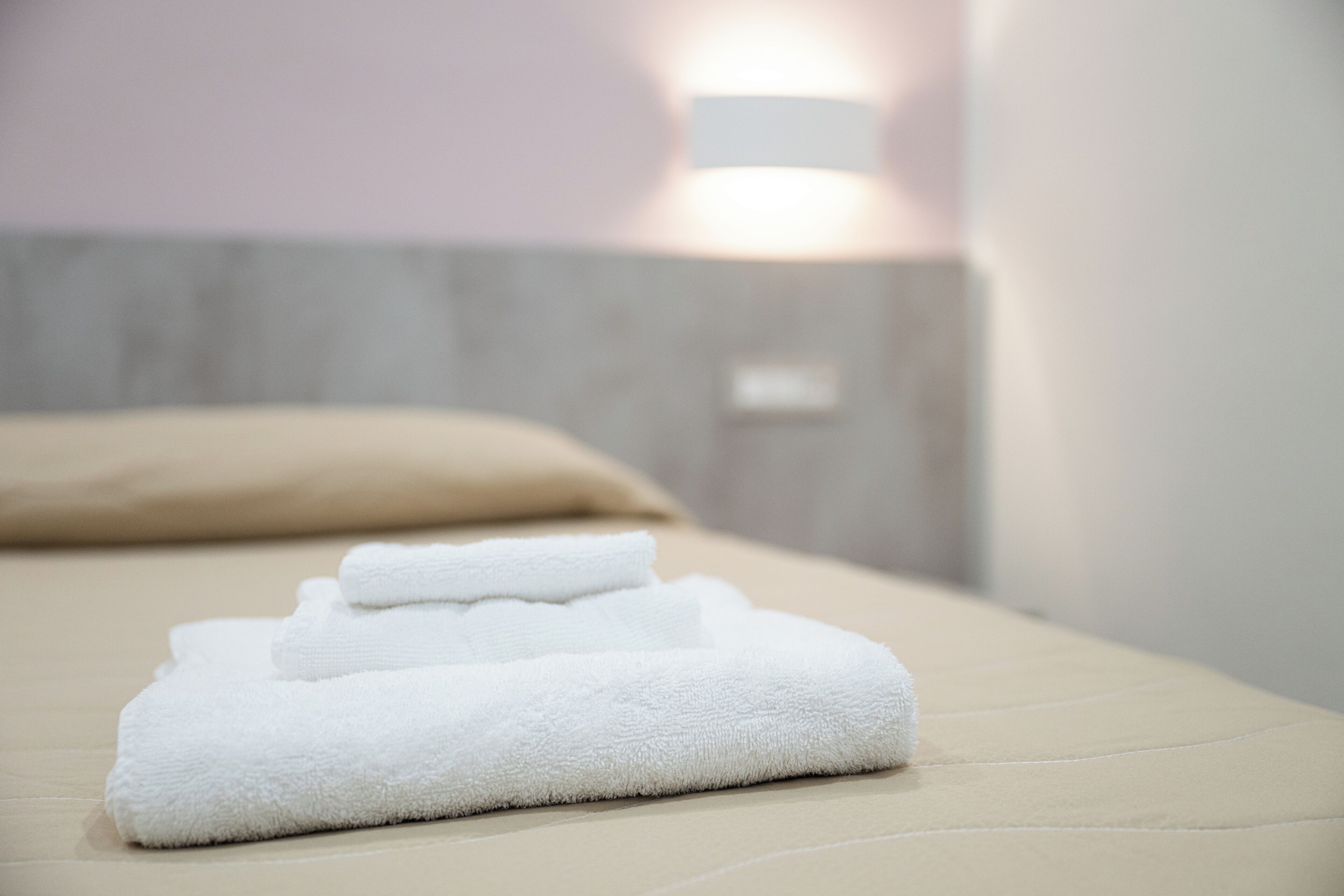 A neatly folded white towel on a bed with a beige headboard and a wall-mounted light fixture.