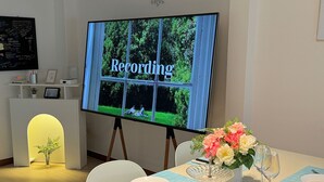Ein Konferenzraum mit einem großen Bildschirm, der 'Aufnahme' anzeigt, einem Tisch gedeckt mit Tellern, Besteck und einer Blumenvase.