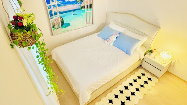 Ein Schlafzimmer mit Bett, Nachttisch, Lampe, Fenster mit Blick auf einen Strand und einer hängenden Pflanze.