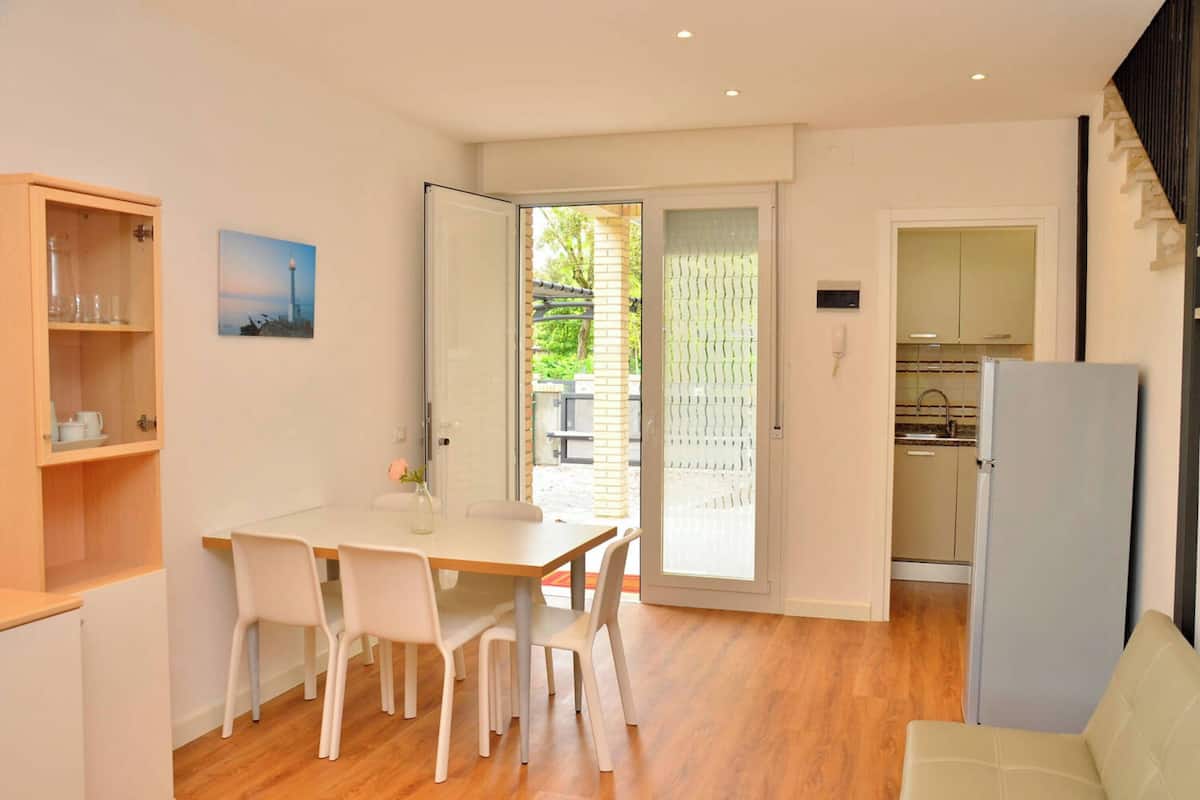 A modern kitchen with a refrigerator, a dining area with a table and chairs, and a view through a glass door to an outdoor area.