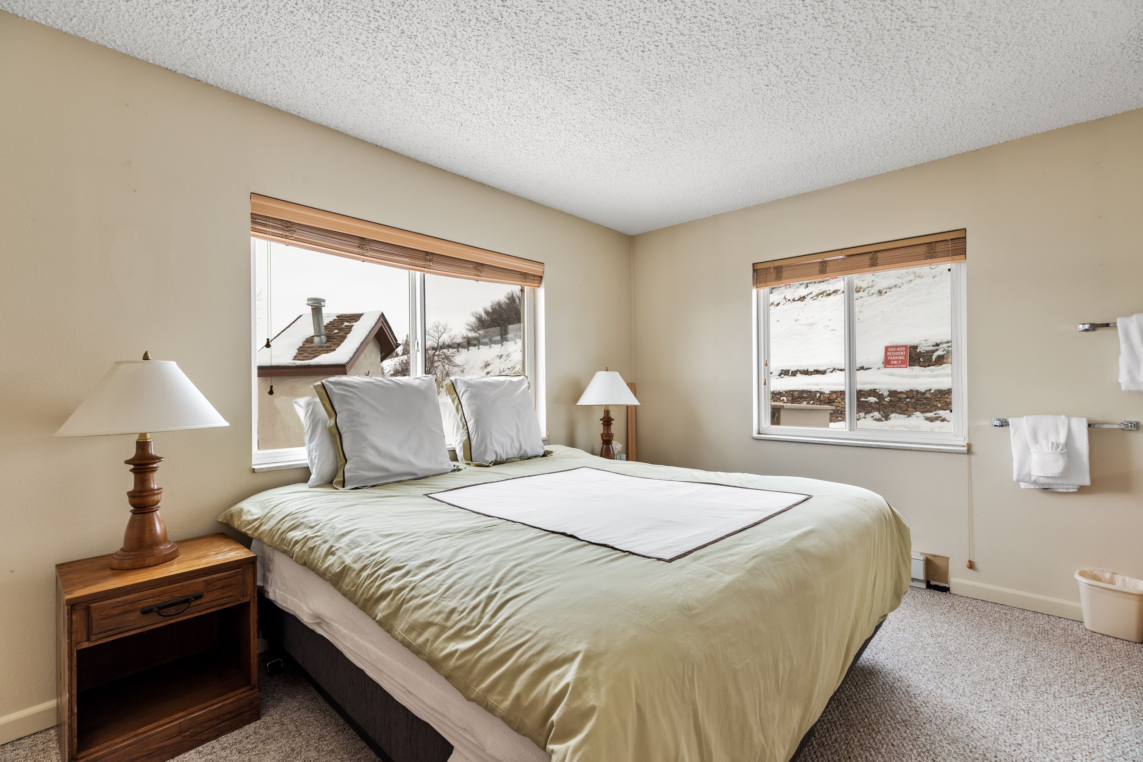A bedroom with a bed, two bedside tables with lamps, a window with a view of a snowy landscape, and a small trash bin.