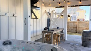 A hot tub with a wooden deck and string lights, a wooden table with stools, and a wooden fence in the background.