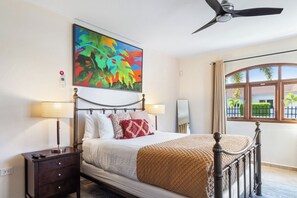 A hotel room with a bed, bedside lamps, a ceiling fan, a window with a view, and a painting on the wall. - Serene 4BR Villa: Pool Views Oasis near Palm Beach (Noord)