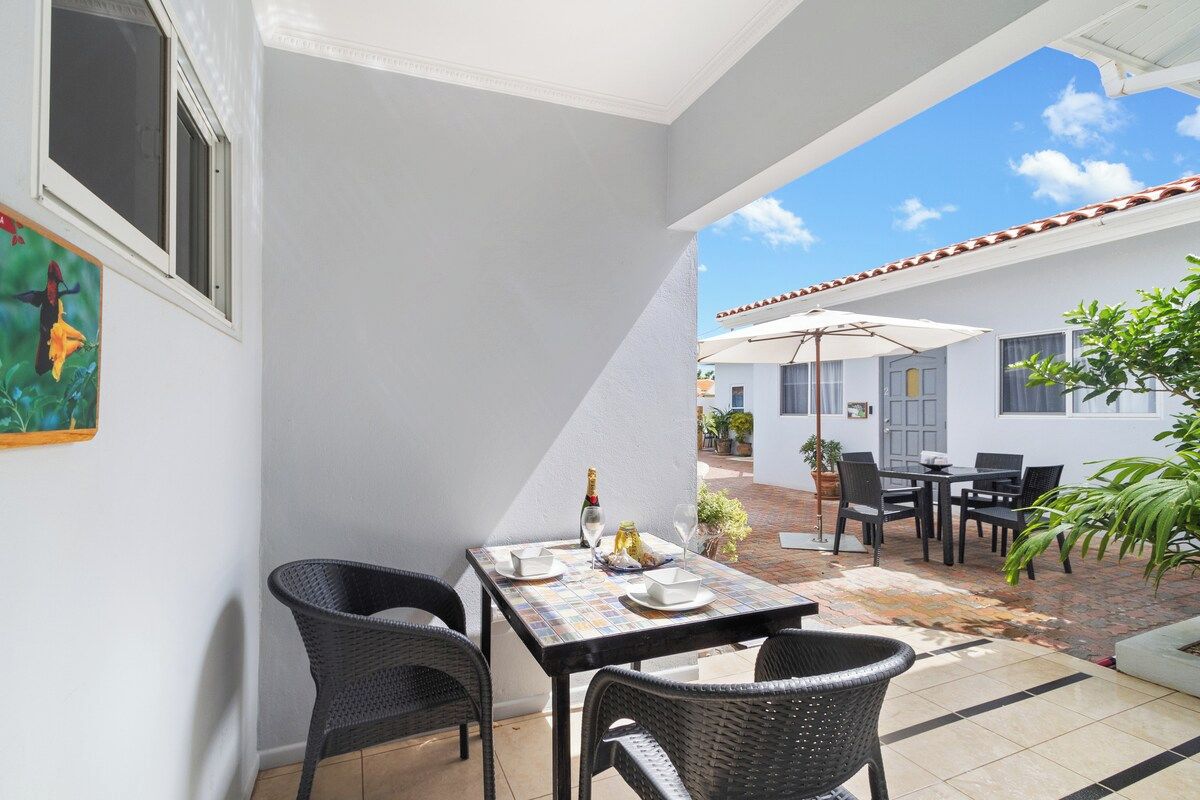 A patio area with a table set for two, surrounded by wicker chairs, a tiled floor, and a view of a white building in the background.
