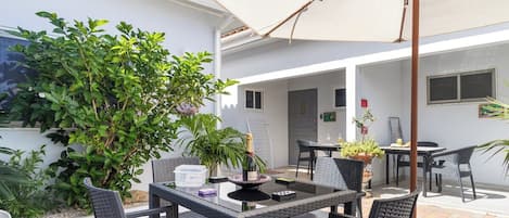 Um pátio externo com mesa e cadeiras, rodeado por plantas em vasos e um guarda-sol grande.
