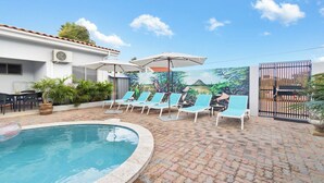 En swimmingpool med liggestole og parasoller, omgivet af en belagt terrasse og et muralmaleri.