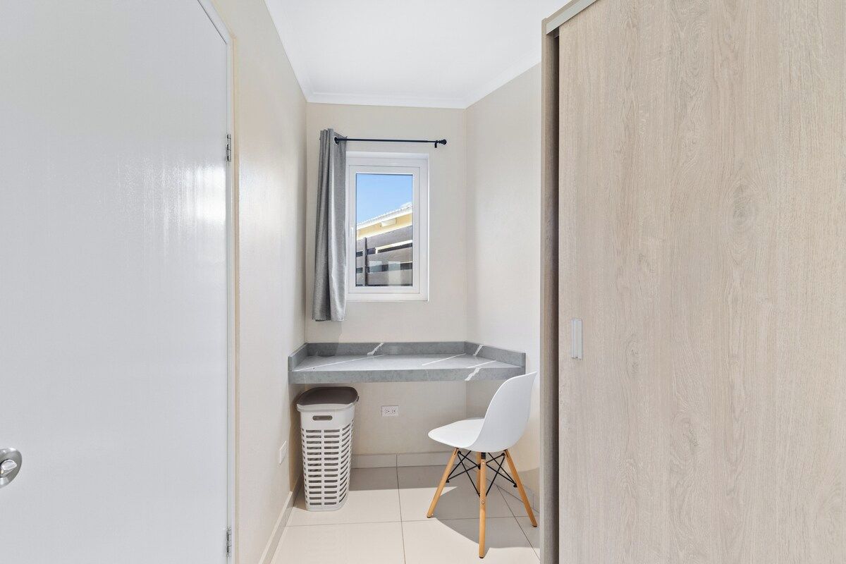 Un baño pequeño con ventana, un perchero, un lavamanos, una silla y un cubo de basura.